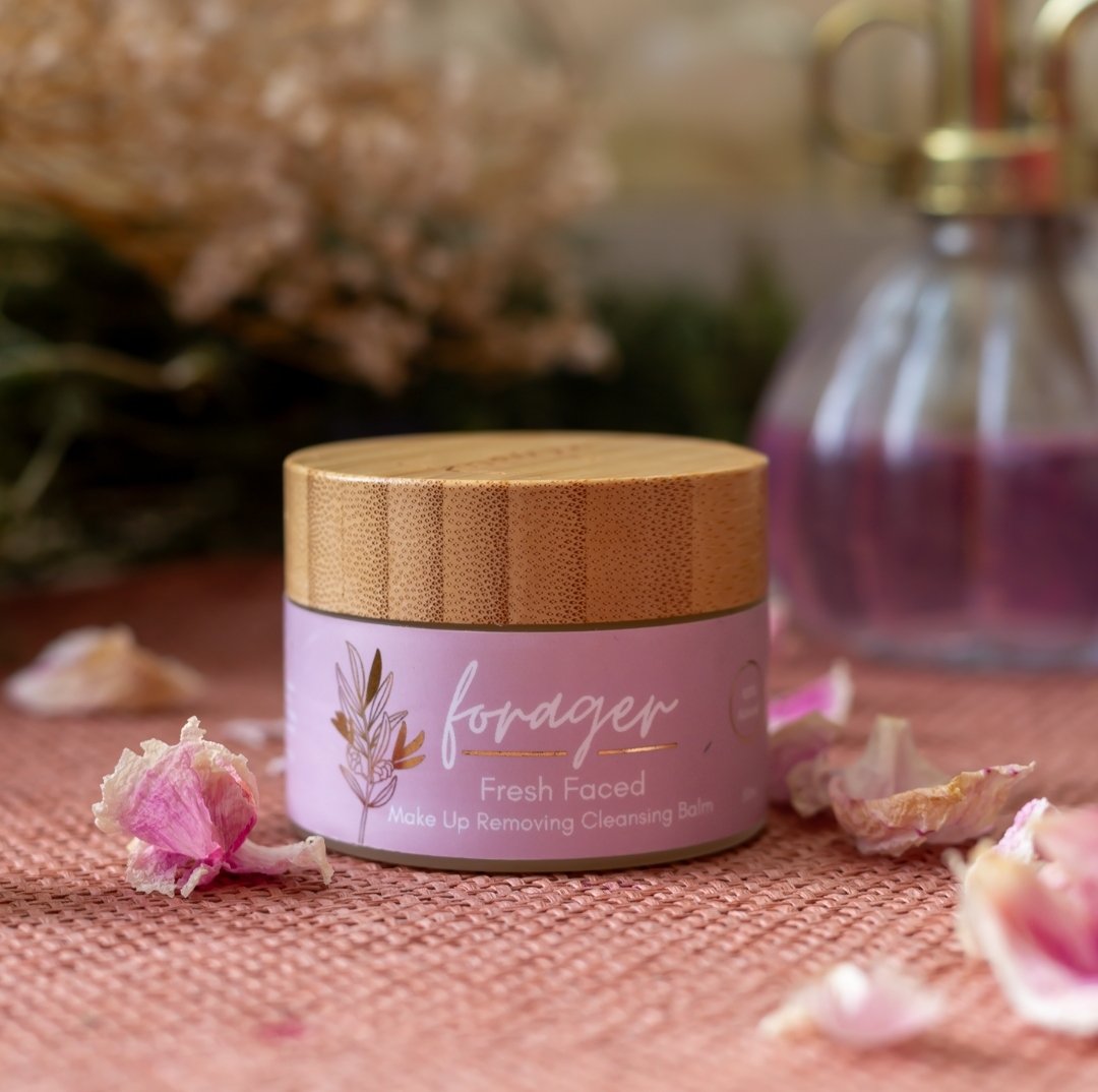 A glass jar with a wooden lid of Forager Fresh Faced Skincare surrounded with pink petals.