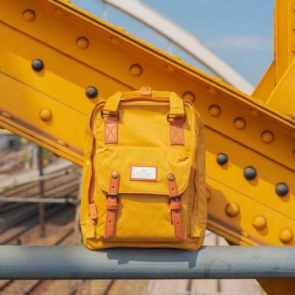 A mustard Doughnut Macaroon backpack sitting on a metal bar leaning against a mustard coloured steel beam.