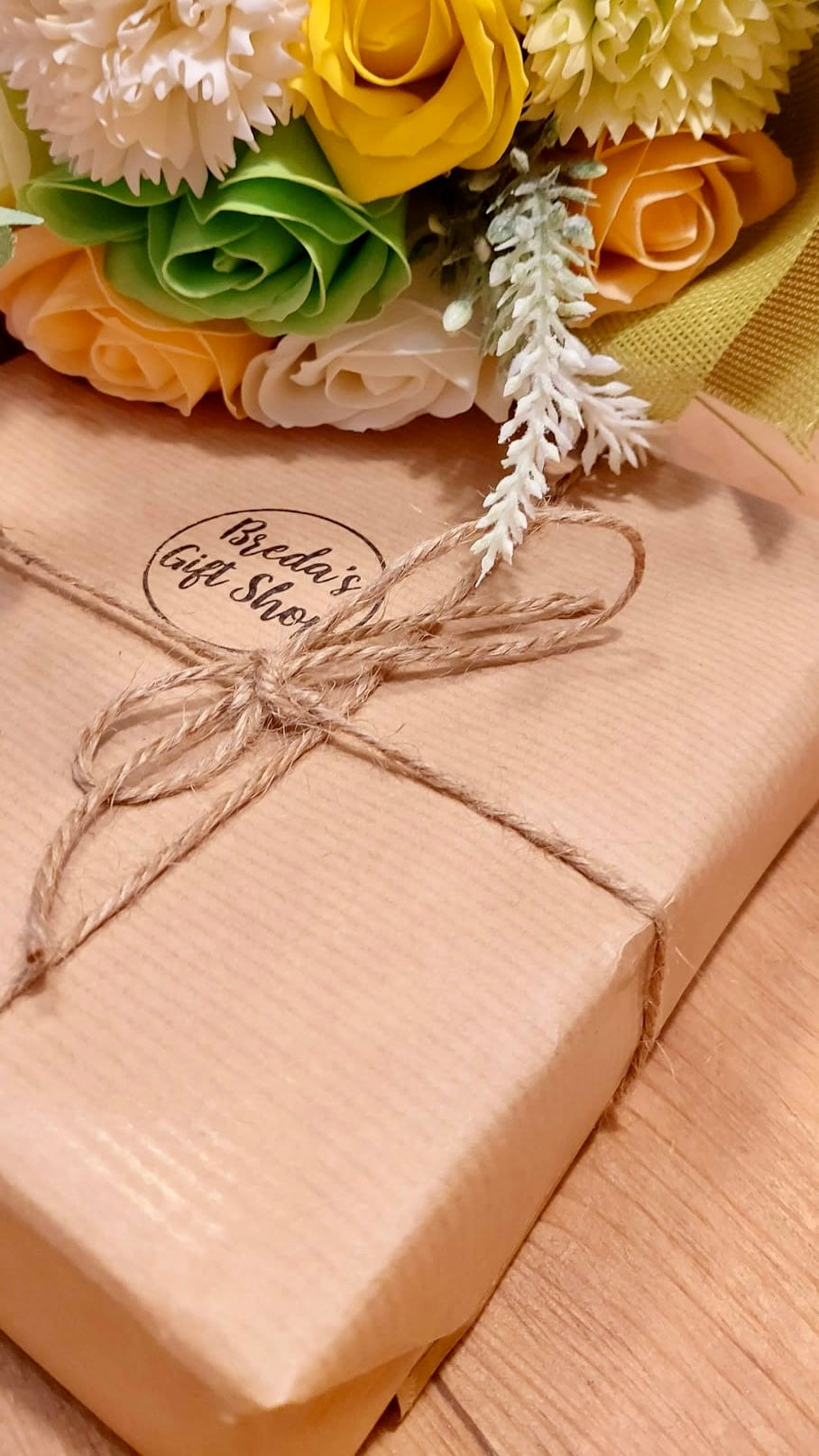 A gift wrapped in brown Kraft paper and twine with a bouquet of soap flowers laying on top of the gift.